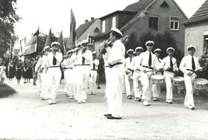 Die schwarz-weiß Fotografie zeigt einen Spielmannszug, der auf einer Straße marschiert. Im Hintergrund sieht man Menschen, die Fahnen führen.