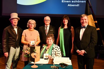 Auf einem Gruppenfoto sieht man drei Männer und drei Frauen, die festlich gekleidet sind und in die Kamera lachen.