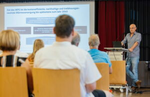 In einem großen Raum sitzen Menschen auf Stuhlreihen. Am rechten Bildrand sieht man einen Mann hinter einem Stehpult, der einen Vortrag hält.