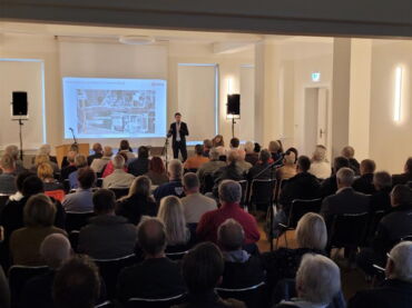 Das Foto zeigte viele Stuhlreihen, auf denen Büger sitzen und einem Vortrag zuhören.