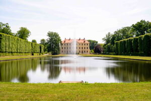Auf einer großen rechteckigen Wasserfläche sieht man mittig eine Fontaine sprudeln. Links und rechts nsieht man in Form geschnittene Bäume. Mittig im Bild steht ein dreistöckiges historisches Gebäude mit vielen Fenstern.