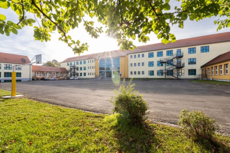 Das Foto zeigt ein farbiges, dreistöckiges Schulgebäude. Davor sieht man einen Schulhof.