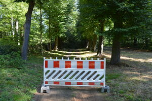 Das Foto zeigt eine rot-weiße Warnbarke, die quer über einen Parkweg gestellt ist. Dahinter sieht man einen Park.