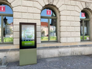 Vor einem historischen Gebäude steht eine ca. 2 Meter hoge Stele. Der untere teil ist grün und im oberen Teil ist ein Fernseher angebracht. Auf diesem sind verschiedene Bilder zu sehen.