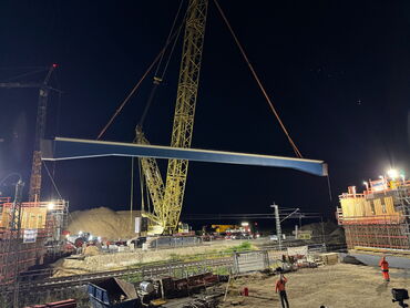 Es ist Nacht. Auf einer hell beleuchteten Baustelle hebt ein riesiger Kran ein meterlanges Stahlstück ein.