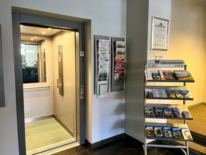 Man sieht einen beleuchteten Fahrstuhl, dessen Tür weit geöffnet ist. Er befindet sich im Rathaus. Man sieht Auslagen für Informationsmaterial rechts neben dem Fahrstuhl.