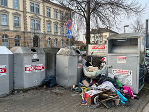 Man sieht einen Platz in der Stadt mit mehreren silbernen Containern. Auf der linken Bildseite sieht man Altglascontainer. Auf der rechten Seite einen Altkleidercontainer, vor dem sich der Müllsammelt.