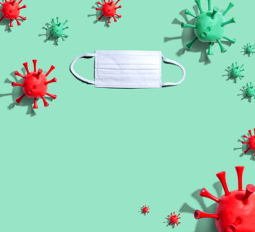 Das Foto zeigt auf grünem Hintergrund grüne und rote Modelle des Corona-Virus und eine OP-Maske.