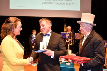 Das Foto zeigt drei Personen, links eine Frau, in der Mitte und rechts je einen Mann. Der Mann in der Mitte übergibt der Frau ein Kästchen in dem eine Medaille liegt.