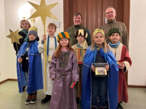 Auf dem Bild sieht man 6 Kinder mit Kronen und Umhängen. Drei Erwachsene stehen hinter ihnen und lächeln in die Kamera.