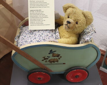 In einem grünen Holz-Puppenwagen mit roten Rädern sitzt ein gelber Plüschteddy. Er hat ein Kissen im Rücken und eine Decke.