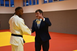 Zwei Männer stehen auf einem gelb-roten Boden. Der Mann rechts im Bild trägt einen Anzug mit Krawatte und hält einen Schlüssel in der Hand. Der Mann links im Bild trägt einen Judoanzug. Beide lächeln.