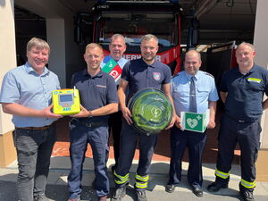 Ein Foto mit sechs Männern, fünf von ihnen tragen Feuerwehruniformen. Sie halten die einzelnen Bestandteile eines Defibrillators in den Händen und lächeln in die Kamera.