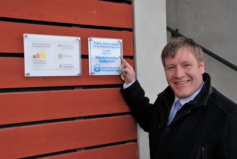 Oberbürgermeister Dr. Sven Mißbach steht neben der neuen Tafel zur Städtebauförderung. Sie hängt neben dem Public Value Award für das öffentliche Bad 2020. Hier hatte das NEB den 2. Platz errungen.