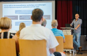 Menschen sitzen auf Stühlen die in Reihen aufgestellt sind. Sie schauen nach vorn auf eine Leinwand. An der rechten Seite des Bildes steht ein Mann ein einem Pult und hält einen Vortrag.