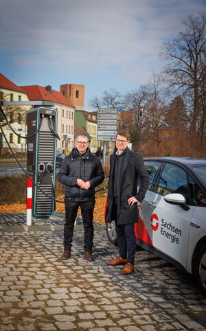 Zwei Männer blicken in die Kamera. Hinter ihnen sieht man eine dunkelgraue Ladesäule für E-Autos. Neben ihnen steht ein weiß-rotes E-Auto.