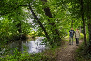 Zwei Menschen, ein Mann und eine Frau, laufen entlang eines Flussufers. Sie tragen Wandersachen und lächeln. Um sie herum sieht man grüne Bäume und Natur.