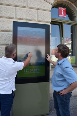 Zwei Männer stehen für eine ca. 2 Meter hohen digiatlen Infowand. Sie schauen sich die Inhalte an. Eine bedient den Tourchsreen.