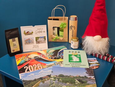 Auf einem Tisch sieht man drapierte Kalender, Bücher, Puzzle und Honig. Daneben sitzt ein Weihnachtsmann.