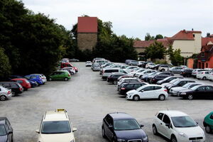 Zu sehen ist ein Platz auf dem Autos parken. Im Hintergrund sind man einen hsitorischen Turm mit Dach. Der Platz wird von Bäumen eingerahmt.