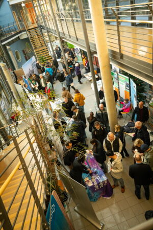 In einem Foyer mit Treppenhaus sieht man unterschiedliche Messestände an denen Personen stehen und reden.