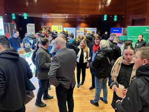 Zu sehen sind viele Menschen, die miteinander sprechen. Manche von ihnen stehen an Messetischen.