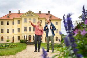 Ein junger Mann und eine junge Frau, beide in Wandersachen, spazieren durch einen Park. Im Hintergrund sind man ein hstorisches Gebäude. Die Frau zeigt dem Mann etwas.