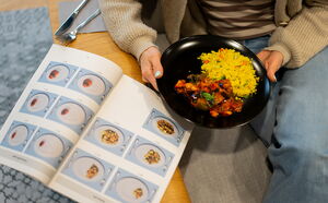 Neben einem aufgeschlagenen Buch mit Fotos von verschiedenen Tellern mit Lebensmitteln in unterschiedlichen Portionsgrößen sitzt eine Frau - man sieht nur ihren Unterkörper - mit einem Teller und vergleicht ihren Teller mit den Fotos im Buch.
