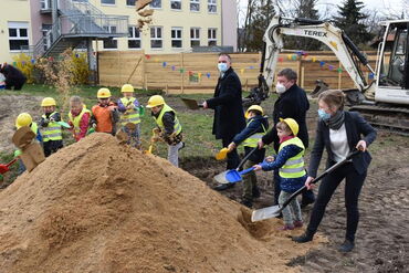 Das Foto zeigt Landrat Ralf Hänsel, Oberbürgermeister Sven Mißbach, Kitaleiterin Kathleen Michel und die Kita-Vorschulkinder wie sie Sand wegschaufeln.