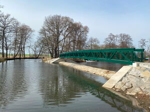 Das Foto zeigt auf der Bildseite einen Fluss und auf der rechten ein neuerrichtetes Wehr. Darauf zu sehen ist eine Brücke mit einem grünen Geländer.