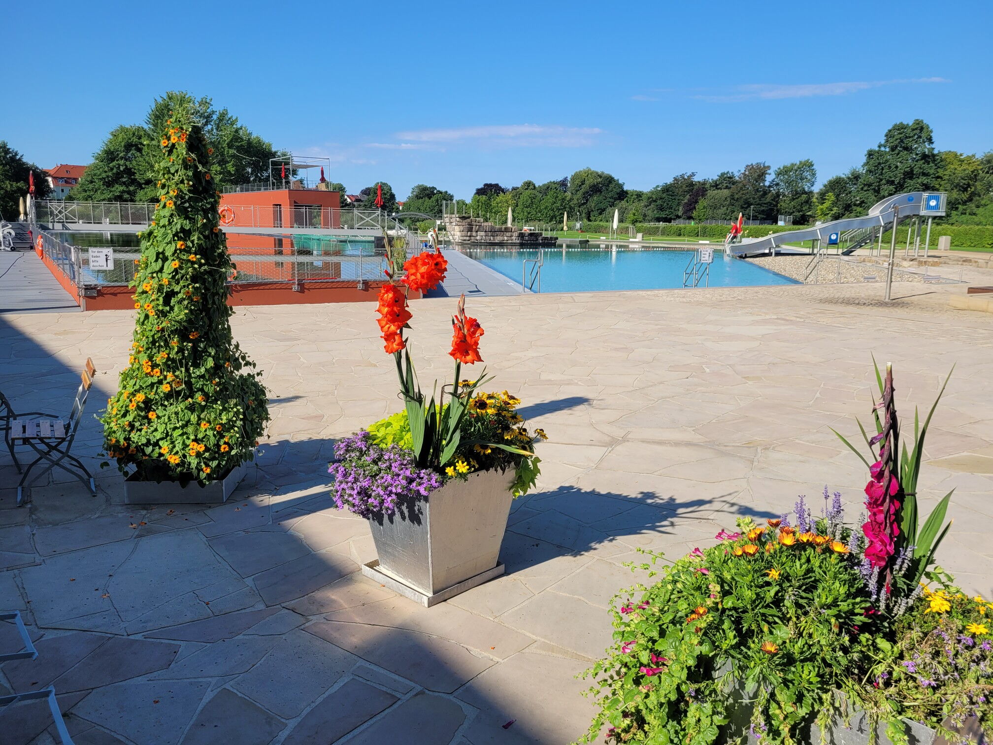 Das Foto zeigt die Außenanlage des NEB. Im Vordergrund sieht man Blumenkübel, dahinter die Wasserfläche mit Rutsche.
