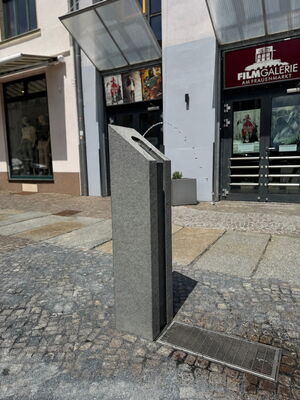 Aus einer grauen Stele fließt ein Wasserstrahl. Im Hintergrund sieht man Gebäude.