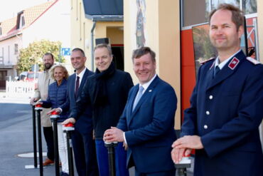 In einer Reihe stehen sechs Menschen, darunter der Landrat und der Oberbürgermeister. Sie lächeln in die Kamera und drücken jeder auf einen roten Knopf. Im Hintergrund sieht man das Feuerwehrgerätehaus in Skaup.